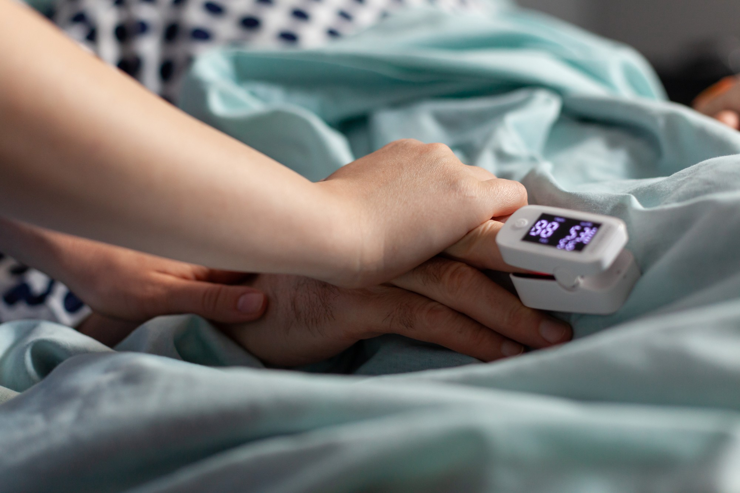 A hand with a pulse oximeter is gently held by another on a bed with teal sheets, conveying a sense of care and concern.