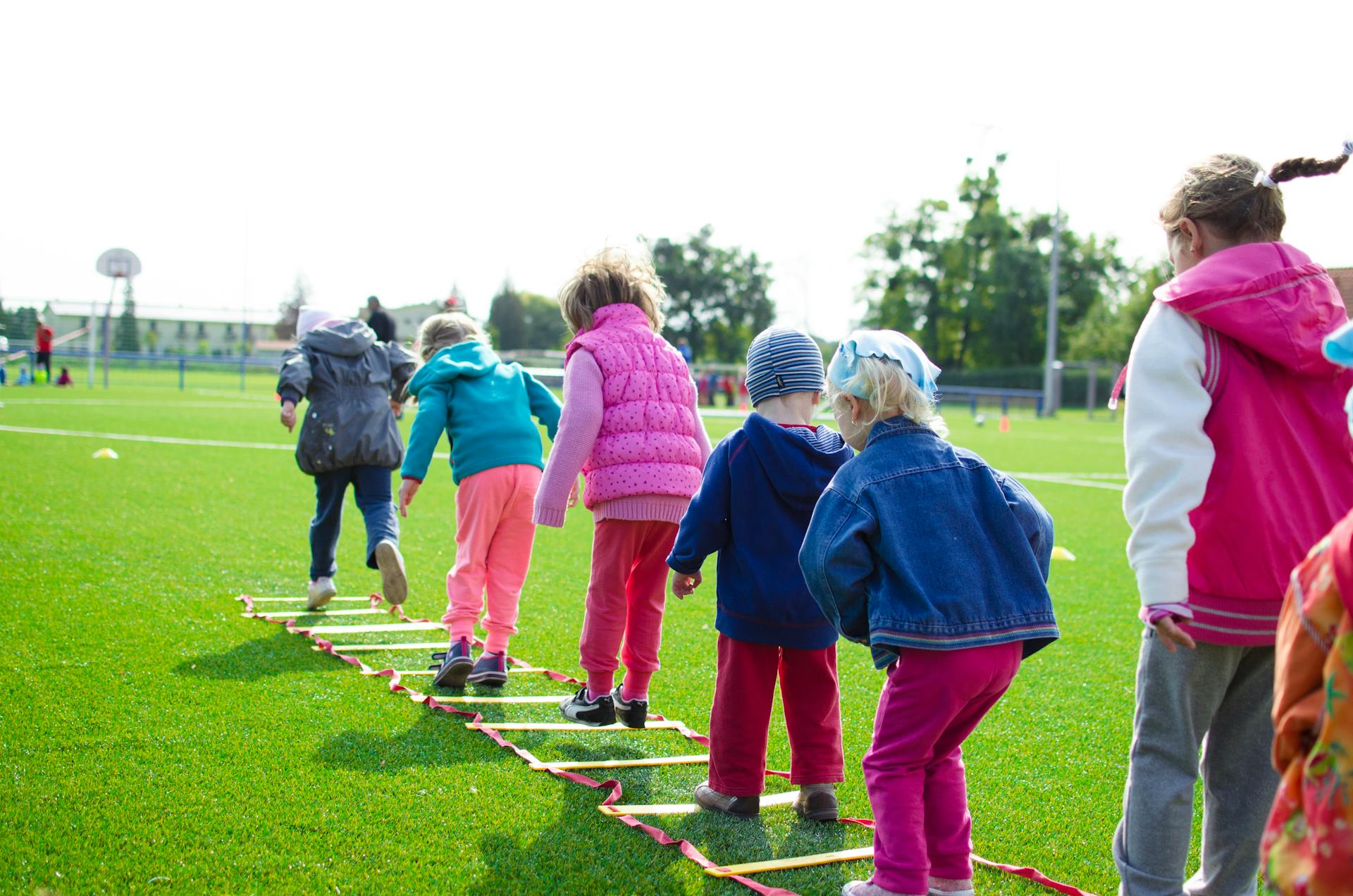 Children in colourful jackets play outdoors, balancing on a ladder laid flat on green grass, showcasing teamwork and fun on a bright day.