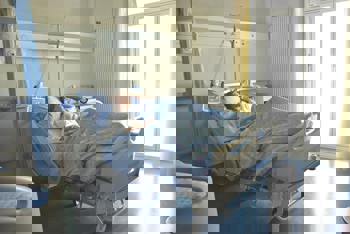 A person rests in a hospital bed, looking contemplative. Sunlight filters through large windows, creating a calm, serene atmosphere in the room.