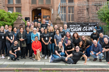 A large group of around 40 people stood outside a brick building, there is a banner which reads "Welcome to the Vision Clinic"