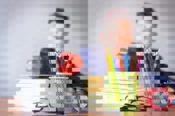 A child sits at a desk with colourful pencils in a green holder, an apple on stacked books, eyeglasses, and a red alarm clock.