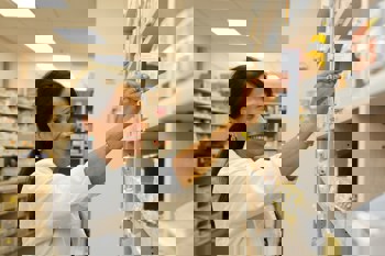 A smiling pharmacist in a white coat selects a medicine box from a pharmacy's organised shelves