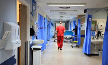 Hospital corridor with blue curtains and medical equipment. A healthcare worker in red scrubs walks down the hallway.