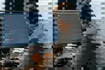 A person in a gray hoodie is focused on typing code on a large monitor. Headphones are placed nearby.