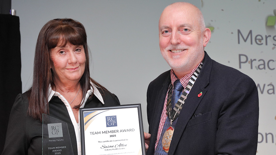 A woman and a man smiling while holding a framed Team Member Award. The woman also holds a trophy. Professional attire; celebratory mood.