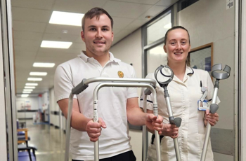 Two healthcare workers in uniform, smiling in a clinic hallway. One holds a walker, the other crutches. 