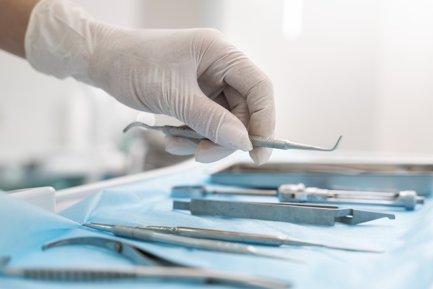 A gloved hand selects a dental tool from an array of instruments on a blue sterile cloth. The setting is a bright, clean dental clinic.
