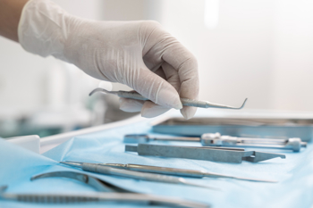 A gloved hand selects a dental tool from an array of instruments on a blue sterile cloth. The setting is a bright, clean dental clinic.