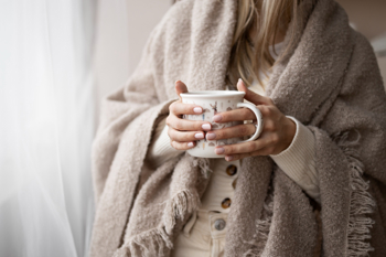 A woman wrapped in a cozy beige blanket holds a patterned mug with both hands. 