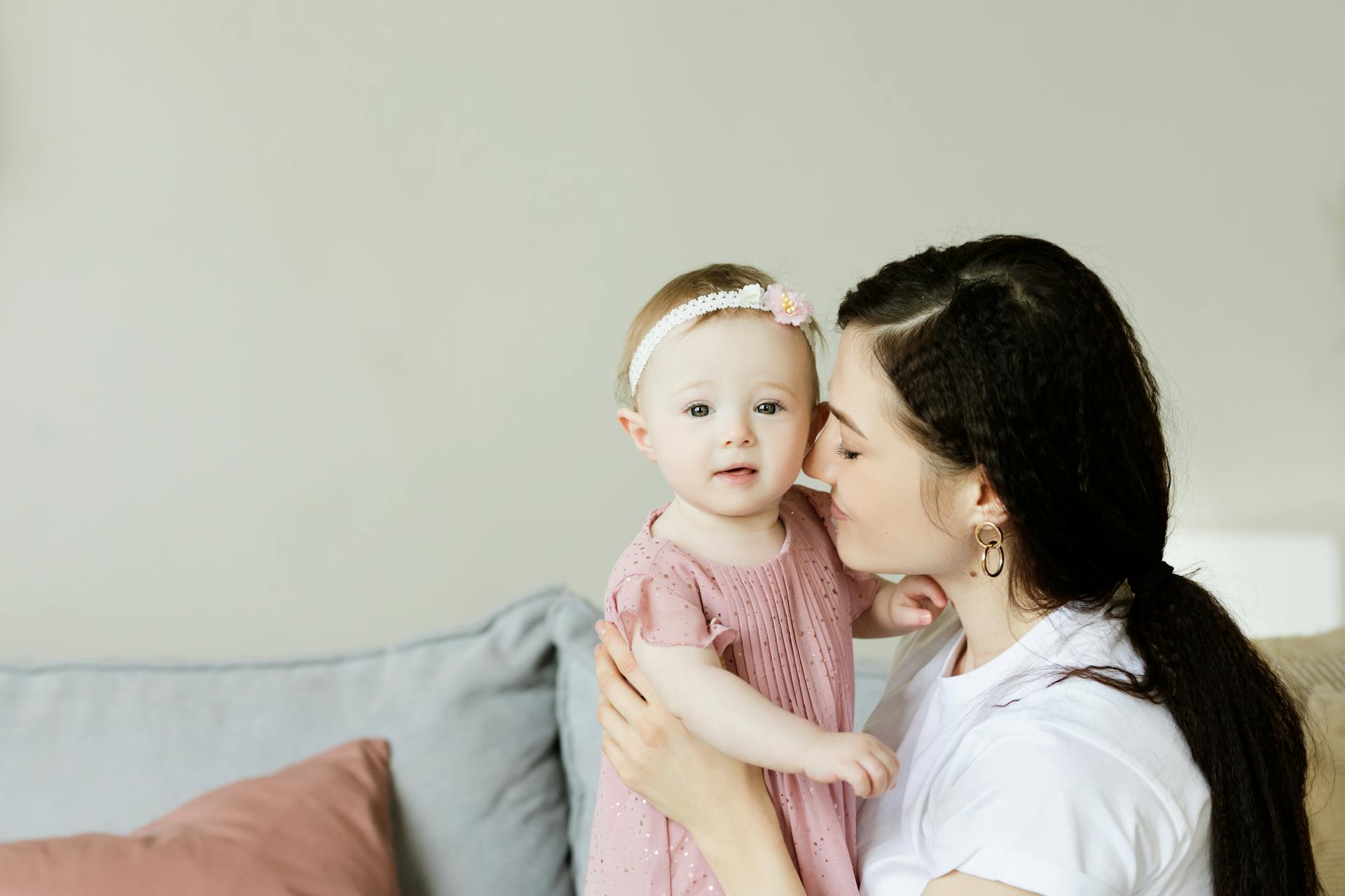 A woman gently kisses a baby on the cheek. The baby wears a pink dress and headband, while the woman is in a white shirt. 